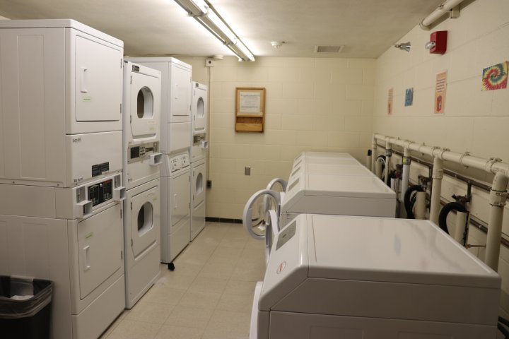 A dormitory laundry room with several stacked washers and dryers along both sides of the room. The space is lit by fluorescent lights and has tiled flooring, with pipes visible on one wall.